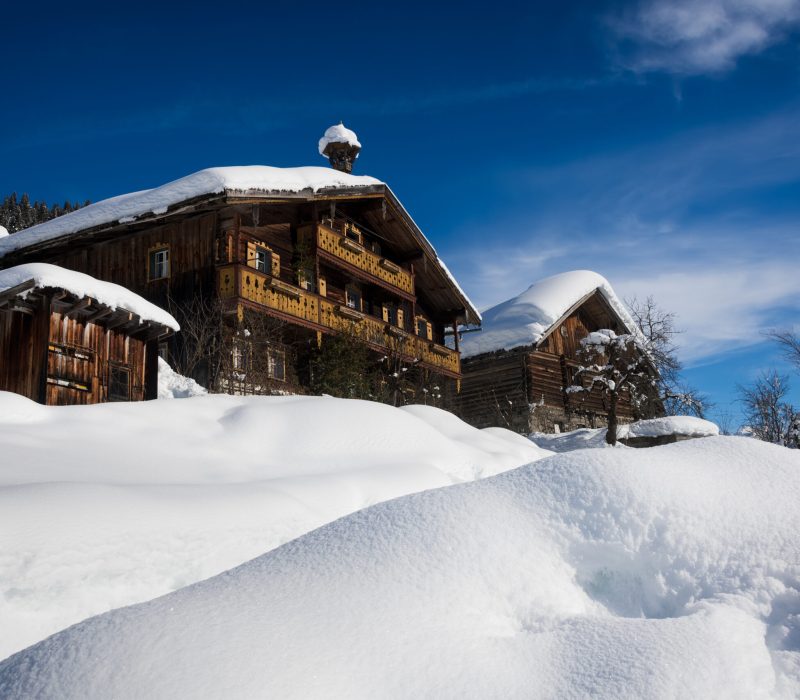 Two wooden ski chalets and a small barn surrounded by and covered with snow under a blue sky in winter in a ski resort in Tyrol, Austria.